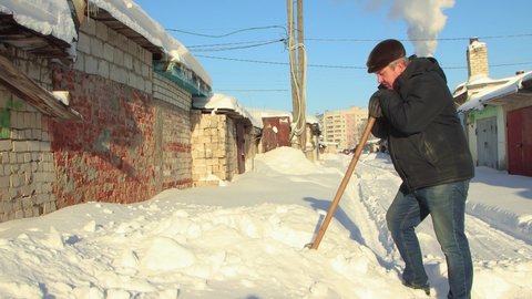 A man stands leaning on a wooden shovel, he cleans the snow and rests. Clear, sunny cool winter day, blue sky. There are snowdrifts all around. Climate change, city, there was a snowstorm. UHD 4K