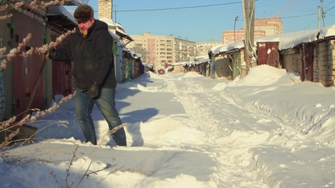 A man cuts large pieces of snow with a shovel and throws them away. Clear, sunny cool winter day, blue sky. There are snowdrifts all around. Climate change, city, there was a snowstorm. UHD 4K