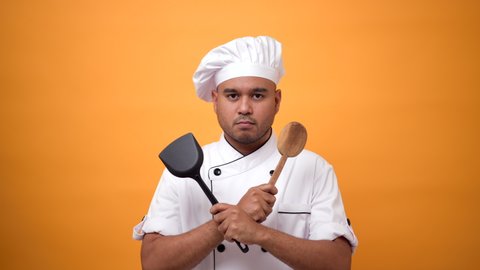 Portrait of man chef holds the kitchen utensils and the turner with a serious expression on isolated yellow background.