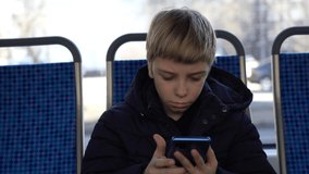 Boy sits at window and looks with interest at screen of smartphone. Happy caucasian kid using cell playing mobile games online on smartphone connected to public wifi sitting on seat in city tram.