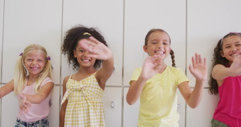 Video of happy diverse girls waving at camera at school corridor. primary school education, learning and socializing concept.
