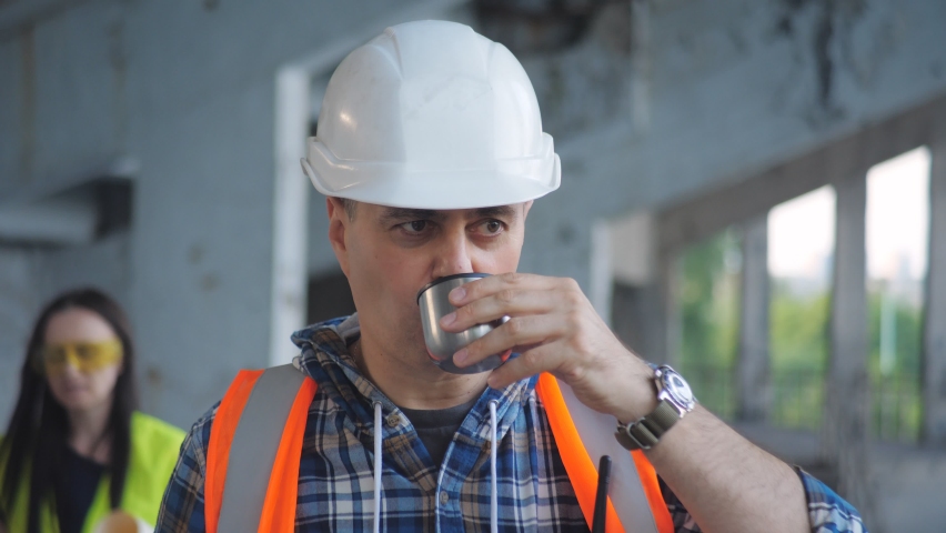 A foreman in a hard hat drinks coffee from a thermos on a construction site during a break, greeting passing employees.