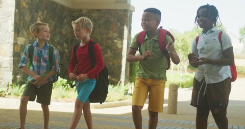 Video of happy diverse boys walking and talking in front of school. primary school education, learning and socializing.