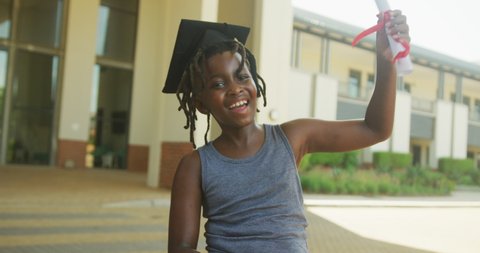 Video of happy african american boy wearing graduation hat and holding diploma in front of school. primary school education, ecology and eco volunteering.