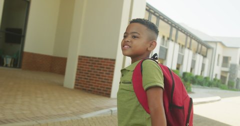 Video of happy african american boy waving to colleagues in front of school. primary school education, learning and socializing.