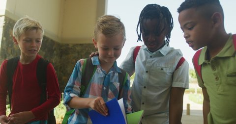 Video of happy diverse boys walking, looking at notebook in front of school. primary school education, learning and socializing.