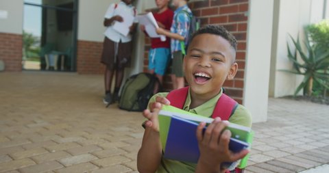 Video of happy african american boy holding books in front of school. primary school education, knowledge and learning concept.
