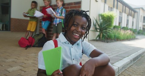 Video of happy african american boy holding books in front of school. primary school education, knowledge and learning concept.