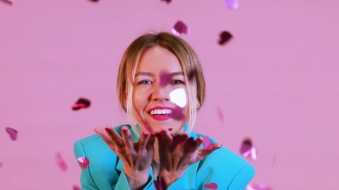 Studio shot of young blond women blowing confetti shape of heart on pink background. Valentines day Birhtday Anniversary 