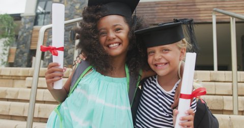 Video of happy diverse girls wearing graduation hats and holding diplomas. primary school education and graduation concept.