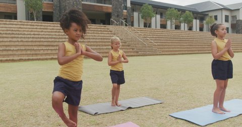 Video of focused diverse girls practicing yoga on mats in front of school. primary school education, sport and exercising concept.