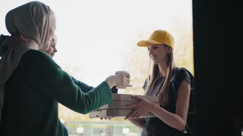 Food delivery. A cheerful courier brings boxes of pizza and drinks, smiles at a Muslim woman and her child at the door of her house, shows pizza. Cafe and restaurant Delivery service.