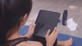 Asian woman in sportswear wiping forehead with towel and using digital tablet while resting after workout in home.