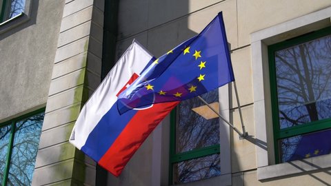 The flags of Slovakia and the European Union flutter in the wind against the background of a wall and large windows in the sunlight