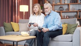 senior couple playing video game console with joysticks sitting on couch in living room. Funny Mature people family having fun spend leisure time together. gray hair man and woman enjoy entertaining 