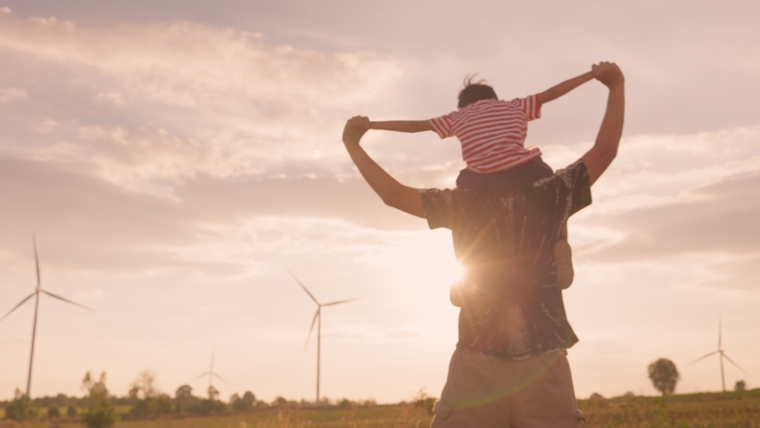 Happy family. An Asian boy sit his father neck and poses for a plane in The windmill farm field producing clean energy. Concept of save the earth and environment