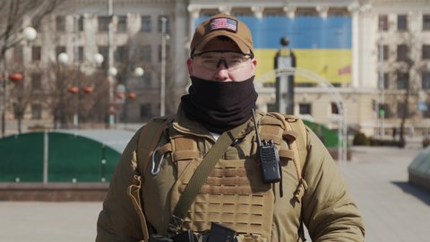 Close-up of a Ukrainian soldier in military uniform and helmet looking at the camera with the flag of Ukraine in the background.
