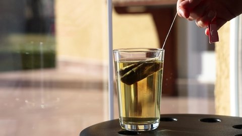Female Hand Brews Herbal Tea in front of Window. Hot Water with Tea Bag in Transparent Glass.