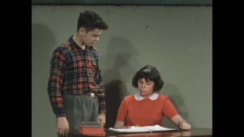 1940s: Girl writes in notebook, looks up and speaks to boy looking over her shoulder. Hand and pen correct grammar of written note. Other boy stands by desk and reads from book, girl and boy talk.