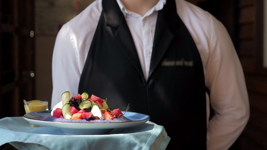 The waiter carries a plate with a delicious salad along the panoramic windows to the client. A waiter in a white shirt and a black apron, serving on the go, in a modern restaurant. Customer service Royalty-Free Stock Footage #1088617401