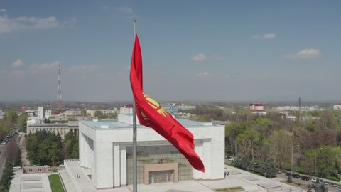 Bushkek, Kyrgyzstan - April 3, 2020: Aerial cinematic footage of red Kyrgyz national flag winding in the wind in golden sun light against panoramic view of centre of Bishkek. Kyrgyzstan symbol. 