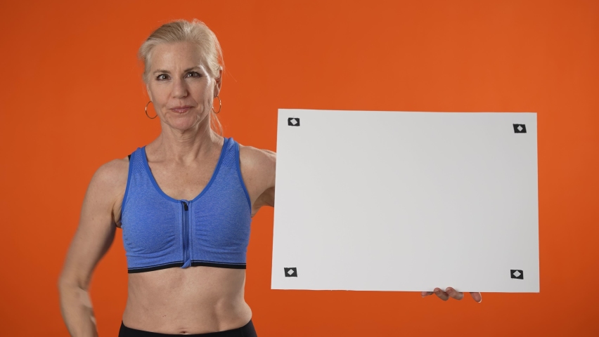 Mature strong sporty athletic fitness woman hold white blank sign board with place for text show muscles isolated on plain orange background
