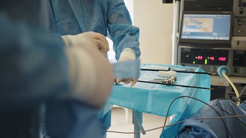 Preparation for surgery. Close up of blue table with laparoscopic instruments, scissors, trocars and cannulas. Close up of male hands in white sterile gloves holding laparoscopic instrument.