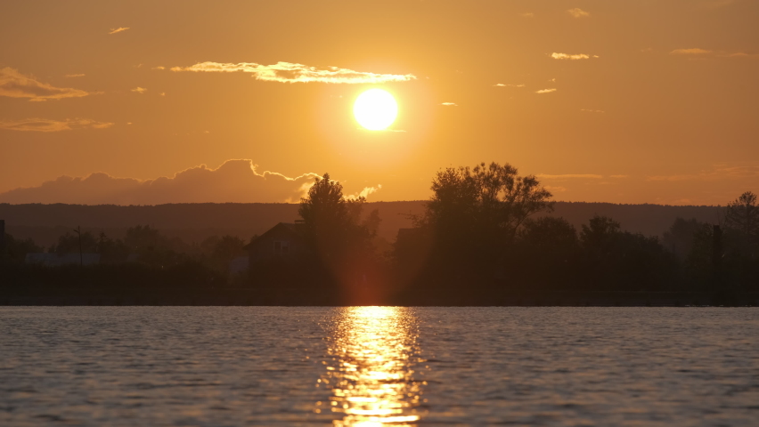 Lakeside landscape with dark silhouette of park trees reflicted in lake water and distant walking pedestrian people on embankment at bright sunset
