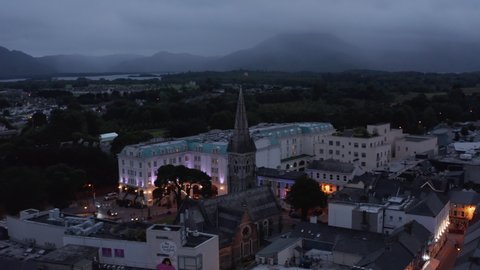 Historic stone church with tall tower and luxury hotel building with illuminated facade. Traffic in evening streets of town. Killarney, Ireland