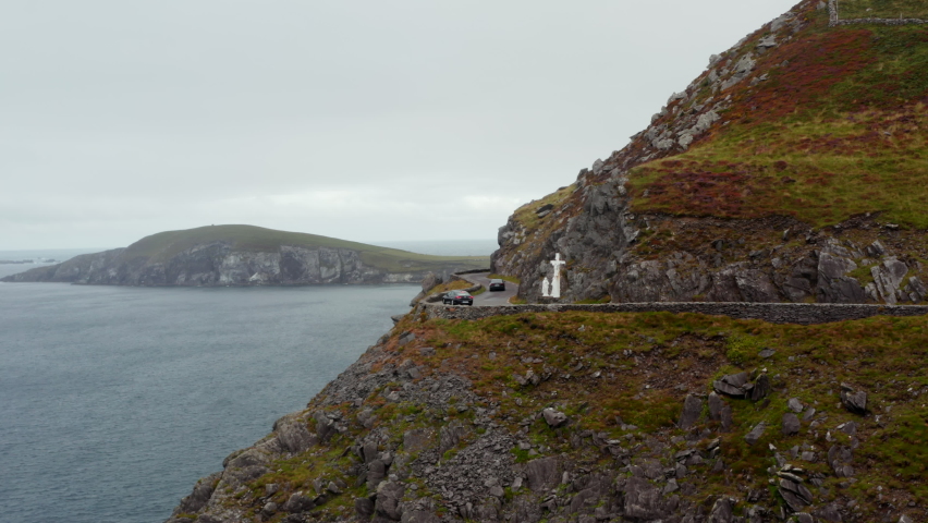 Car waiting in curve on oncoming vehicle. Difficult driving on panoramic coastal route. White sculpture near road, Cross at Slea Head. Ireland