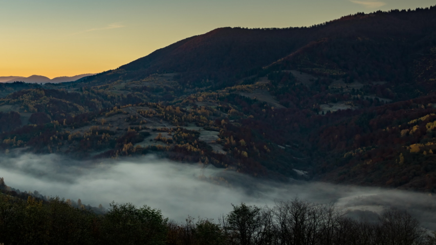 Sun rises in highland with big forestry mountains and morning fog fills valley among hill slopes in autumn under orange sky 4K time-lapse video
