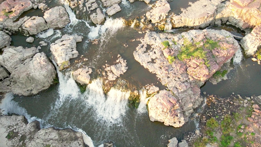 Overhead view of water falls in Falls Park, Sioux Falls, South Dakota. Water taking different paths through the landscape with calmer ponds spread out. Sun reflecting off the water. 