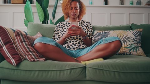 Happy young African American woman chatting with friends using application in phone and smiling sending SMS message in dating app sits cross-legged on sofa in living room. Lifestyle, recreation