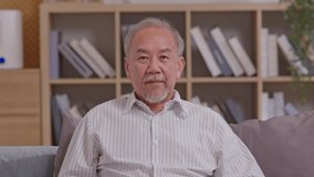 Grey haired senior asian man wearing casual white shirt sitting on couch waving saying hello happy and smiling in video conference call, friendly welcome gesture posture. Comfy and relax at home.