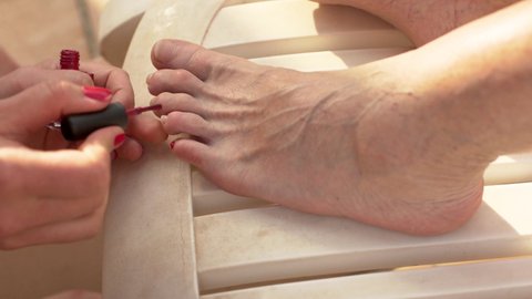 Young woman applying red nail polish on older woman feet, relaxing at plastic sunbed, closeup detail