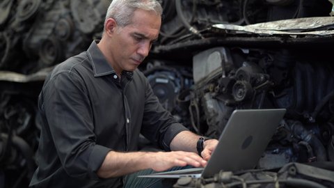 senior engineer using laptop computer checking list old used car engine in machinery parts shop warehouse. old technician man Repair vehicle automobile machine in Recycle motor factory