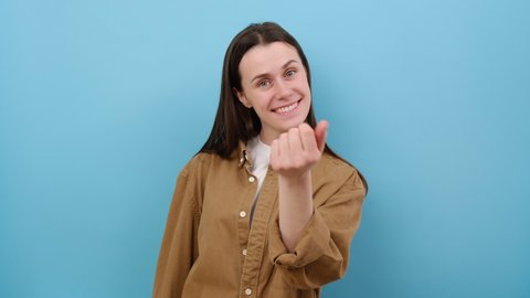 Come here. Portrait of attractive young woman making beckoning gesture, inviting to come, flirting and looking playful, wearing brown shirt, posing isolated over blue color background wall in studio