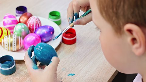 A boy painting an Easter egg with blue paintbrush on a wooden table. Colorful easter eggs. Easter Holidays preparation theme. Selective focus