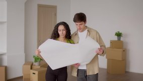 Young Spouses Holding Paper With Renovation Plan Standing Among Moving Boxes In Their New Home. Real Estate Ownership And Purchase, Family Housing Concept. Slow Motion