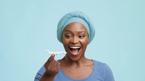 Travel Concept. Cheerful African American Lady Playing With Plane Toy Model Planning Vacation Smiling To Camera Standing Over Blue Studio Background, Wearing Headwrap. Traveling And Transportation