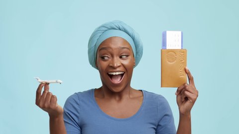 Vacation. Excited African American Female Holding Travel Tickets And Plane Toy Model Smiling To Camera Standing Over Blue Studio Background, Wearing Headwrap. Traveling Concept