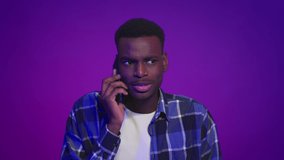 African American Guy Talking On Cellphone Looking At Camera Standing Over Purple Studio Background. Millennial Man Chatting On Phone. Mobile Communication Concept. Slowmo