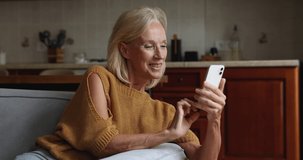 Happy middle-aged woman staring at smartphone screen, text message, share sms to friend, enjoy remote communication by cell sit on cozy sofa at home. Modern tech usage, fun, mobile apps usage concept