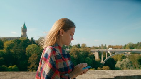 Young European Woman Texting on Mobile App using Smartphone in Luxembourg City Walking near Landmark Adolphe Bridge. Tourist, Student or Business Lady uses Phone. 4K medium tracking shot