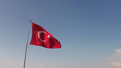 A large bright flag of Turkey flutters on a flagpole high above the ground against the backdrop of the sea, mountains and clouds. Aerial 4k shooting from a drone