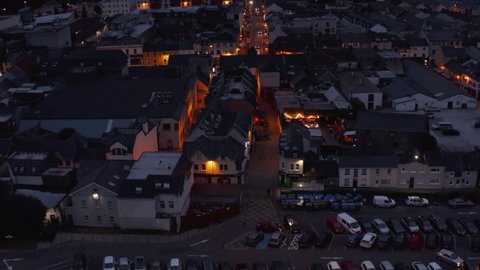 Forwards fly above illuminated street in town after sunset. Tilt up reveal of overcast sky and mountains in distance. Killarney, Ireland