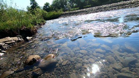 Camera descends into the water of shallow mountain river. Moving glare of sun on surface of rocks. Turbid muddy water. Underwater view with stones covered mule at bottom. Flow of water in river inside