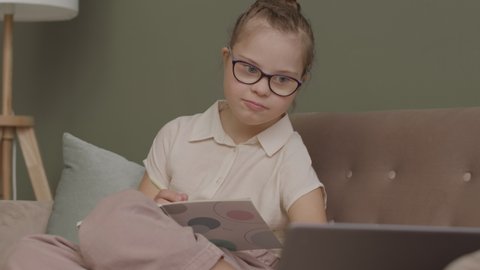 Slowmo shot of pretty 11-year-old girl in eyeglasses sitting on sofa at home and having online lesson on laptop