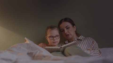 Slowmo shot of mother reading book to her little daughter at night, lying together in bed at cozy girls bedroom