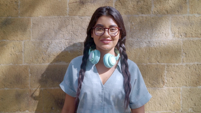 A smiling young Indian Asian hipster modern stylish woman or female is outdoors headphones around neck standing against a wall wearing eye glasses confidently looking in the camera. Retro fashion Royalty-Free Stock Footage #1088659317